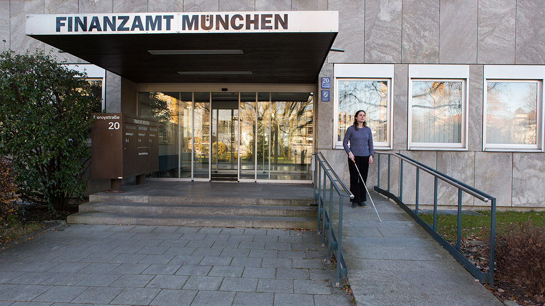 Eingang des Finanzamts München. Eine Frau mit Langstock geht eine Rampe hinunter.