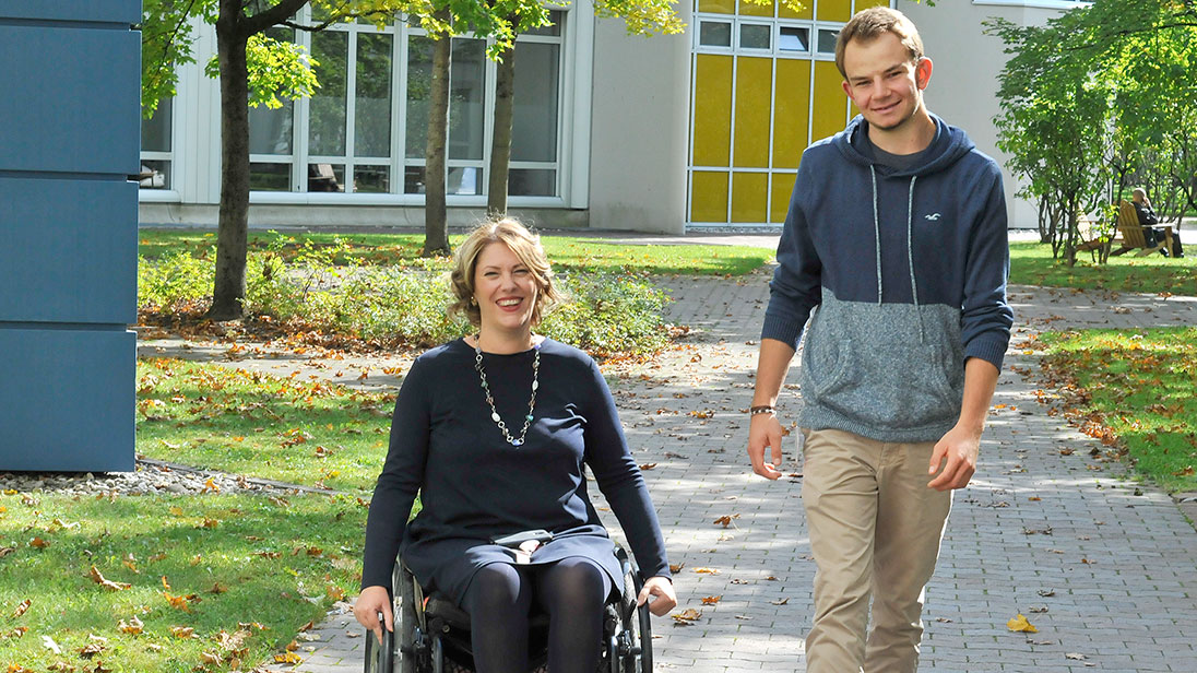 Szene im Freien: Frau im Rollstuhl und junger Mann zu Fuß bewegen sich auf die Kamera zu.