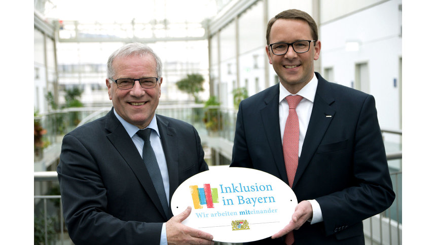 Johannes Hintersberger übergibt das Emblem „Inklusion in Bayern – Wir arbeiten miteinander“ an Dr. Christian Hartel.