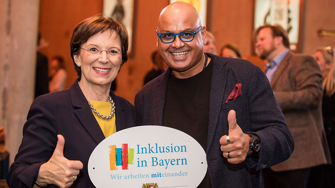 Staatsministerin Emilia Müller und Saliya Kahawatte mit dem Emblem „Inklusion in Bayern – Wir arbeiten miteinander“.